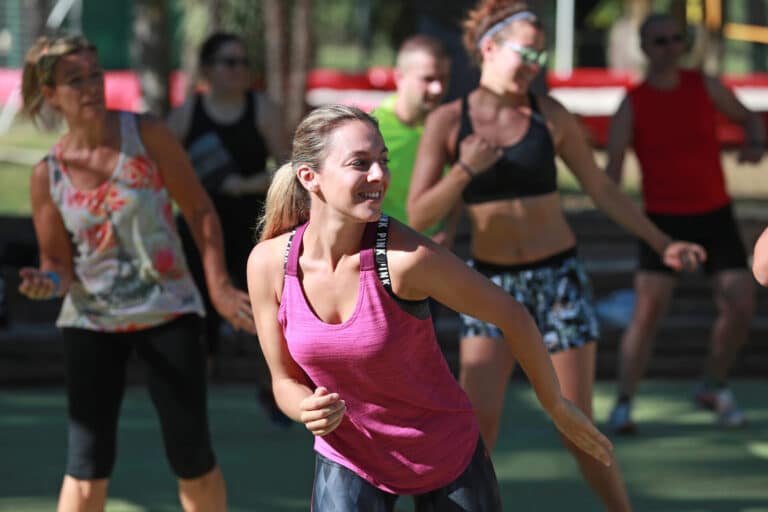 En gruppe mennesker, der deltager i en udendørs fitness- eller dansetime. Fokus er på en kvinde i en lyserød tanktop, der smiler og bevæger sig energisk. Andre i baggrunden er også engageret i aktiviteten, iklædt afslappet træningstøj.