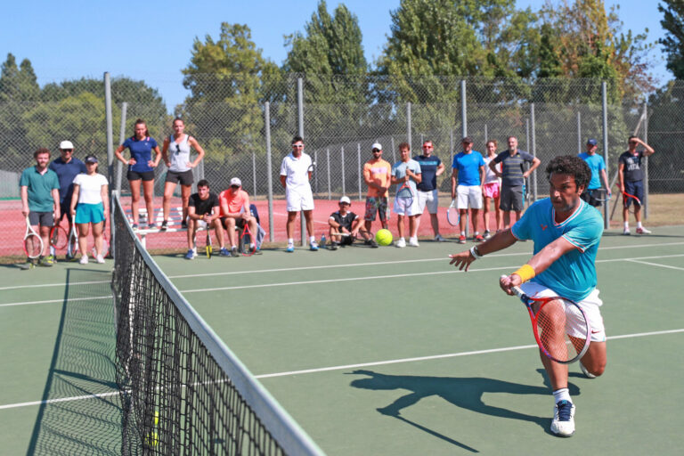 En tennisspiller i blåt og orange slår en forehand nær nettet, mens en gruppe mennesker med ketsjere ser til fra sidelinjen. Scenen foregår på en solskinsdag på en udendørs tennisbane.