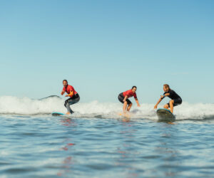 Tre mennesker surfer på små bølger under en klar blå himmel. Hver bærer en våddragt og kører på separate surfbrætter i havet. Det ser ud til at de nyder aktiviteten og viser balance og dygtighed. Vandet er roligt og blåt og afspejler himlen ovenover.