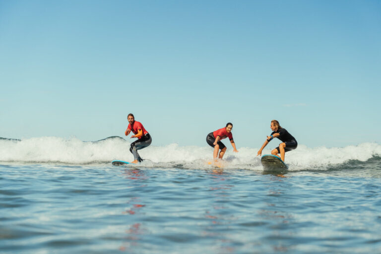 Tre mennesker surfer på små bølger under en klar blå himmel. Hver bærer en våddragt og kører på separate surfbrætter i havet. Det ser ud til at de nyder aktiviteten og viser balance og dygtighed. Vandet er roligt og blåt og afspejler himlen ovenover.