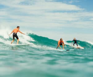 En gruppe mennesker surfer på en blå bølge under en delvist overskyet himmel. Fire surfere iført våddragter rider på bølgen med varierende dygtighed og balance og skaber stænk af vand, mens de bevæger sig. Havet strækker sig til horisonten i baggrunden.