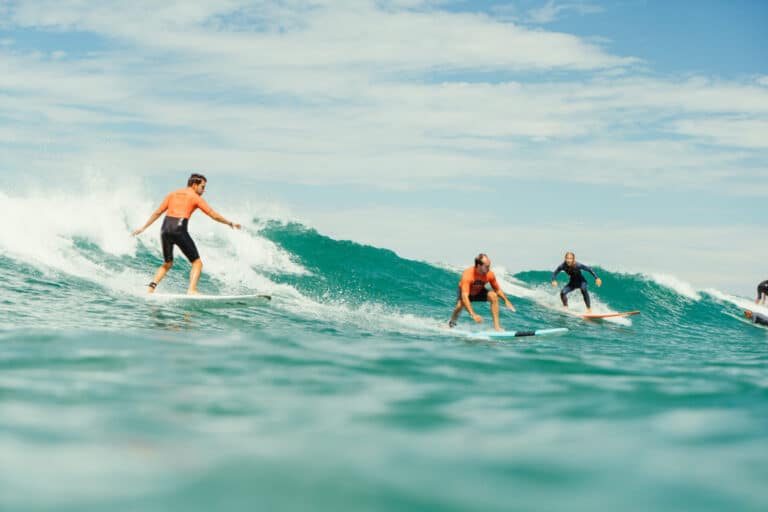 En gruppe mennesker surfer på en blå bølge under en delvist overskyet himmel. Fire surfere iført våddragter rider på bølgen med varierende dygtighed og balance og skaber stænk af vand, mens de bevæger sig. Havet strækker sig til horisonten i baggrunden.