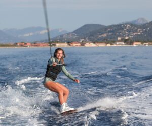 En person, der wakeboarder på et roligt hav nær kysten, iført en redningsvest og holder fast i et slæbetov og blander sport med eventyr. Baggrunden byder på bjerge og en lille by under en klar himmel, perfekt til sportsferie-entusiaster, der søger spænding.