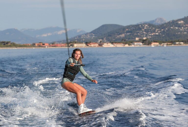 En person, der wakeboarder på et roligt hav nær kysten, iført en redningsvest og holder fast i et slæbetov og blander sport med eventyr. Baggrunden byder på bjerge og en lille by under en klar himmel, perfekt til sportsferie-entusiaster, der søger spænding.