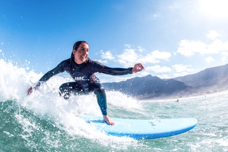 En person surfer på en klar dag og rider på en bølge på et blåt surfbræt. De har en våddragt på og smiler med havet og fjerne bjerge i baggrunden. Himlen er lys med spredte skyer.