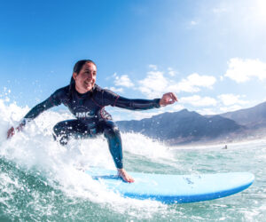 Person, der surfer på et blåt surfbræt, skærer gennem en bølge under en klar blå himmel. De er iført en mørk våddragt og smiler, med bjerge og en sandet kystlinje synlig i baggrunden.