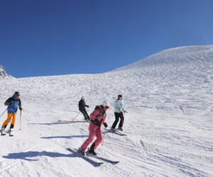 Fire personer på ski ned ad en sneklædt skråning under en klar blå himmel. De er klædt i farverigt skiudstyr med snedækkede bjerge synlige i baggrunden. Terrænet er bredt og åbent, ideelt til skiløb.