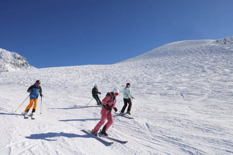 Fire personer på ski ned ad en sneklædt skråning under en klar blå himmel. De er klædt i farverigt skiudstyr med snedækkede bjerge synlige i baggrunden. Terrænet er bredt og åbent, ideelt til skiløb.